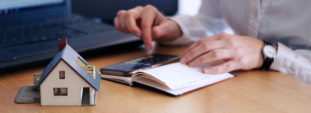 Primer plano de una persona. Manos sobre el escritorio mientras hace cuentas con una calculadora. Sobre el escritorio se encuentra la maqueta de una casa. Al fondo, una computadora portátil.
